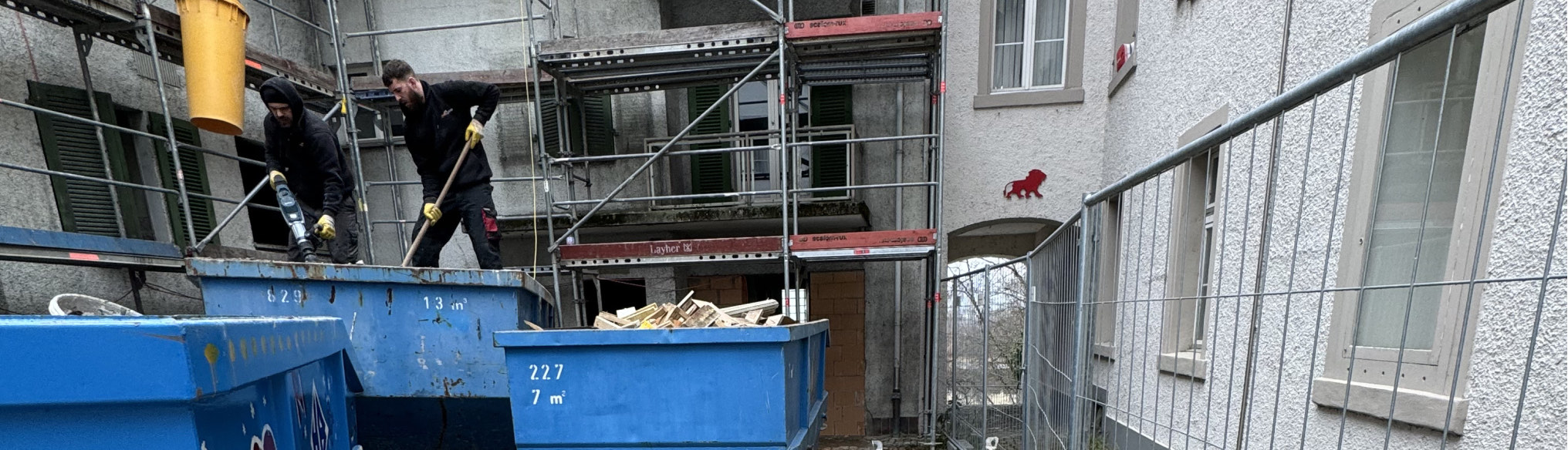 Arbeiter füllen Mulden vor einem eingerüsteten Haus